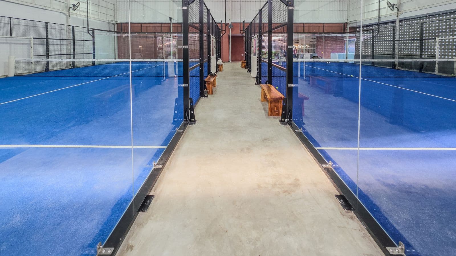Two glass-walled indoor padel courts either side of a walkway at a padel club