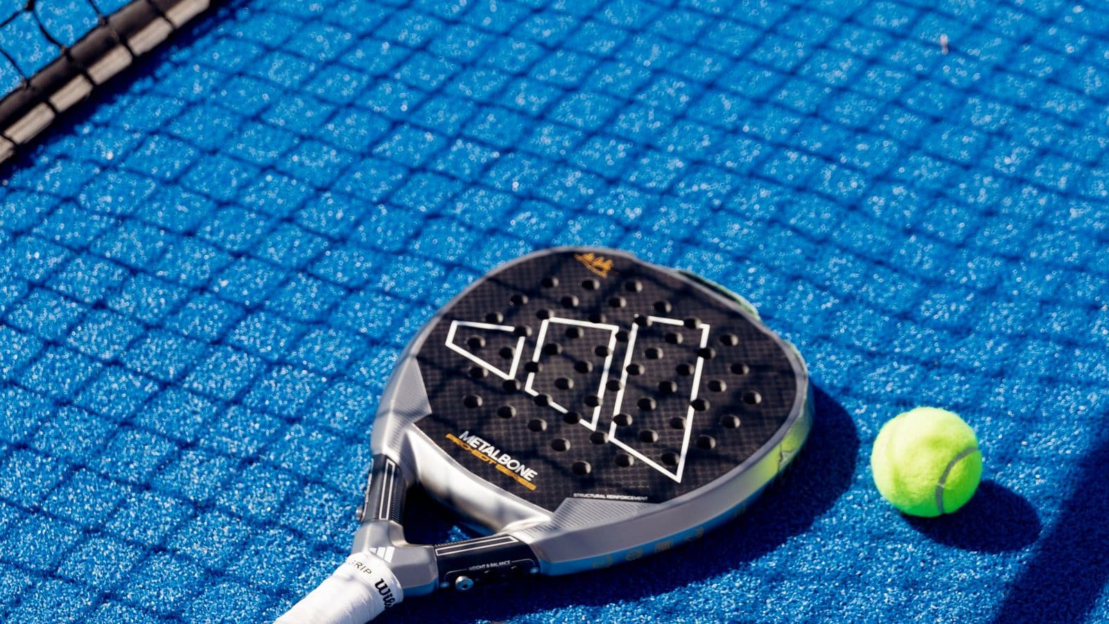A padel racket and ball resting on blue court turf beside the net